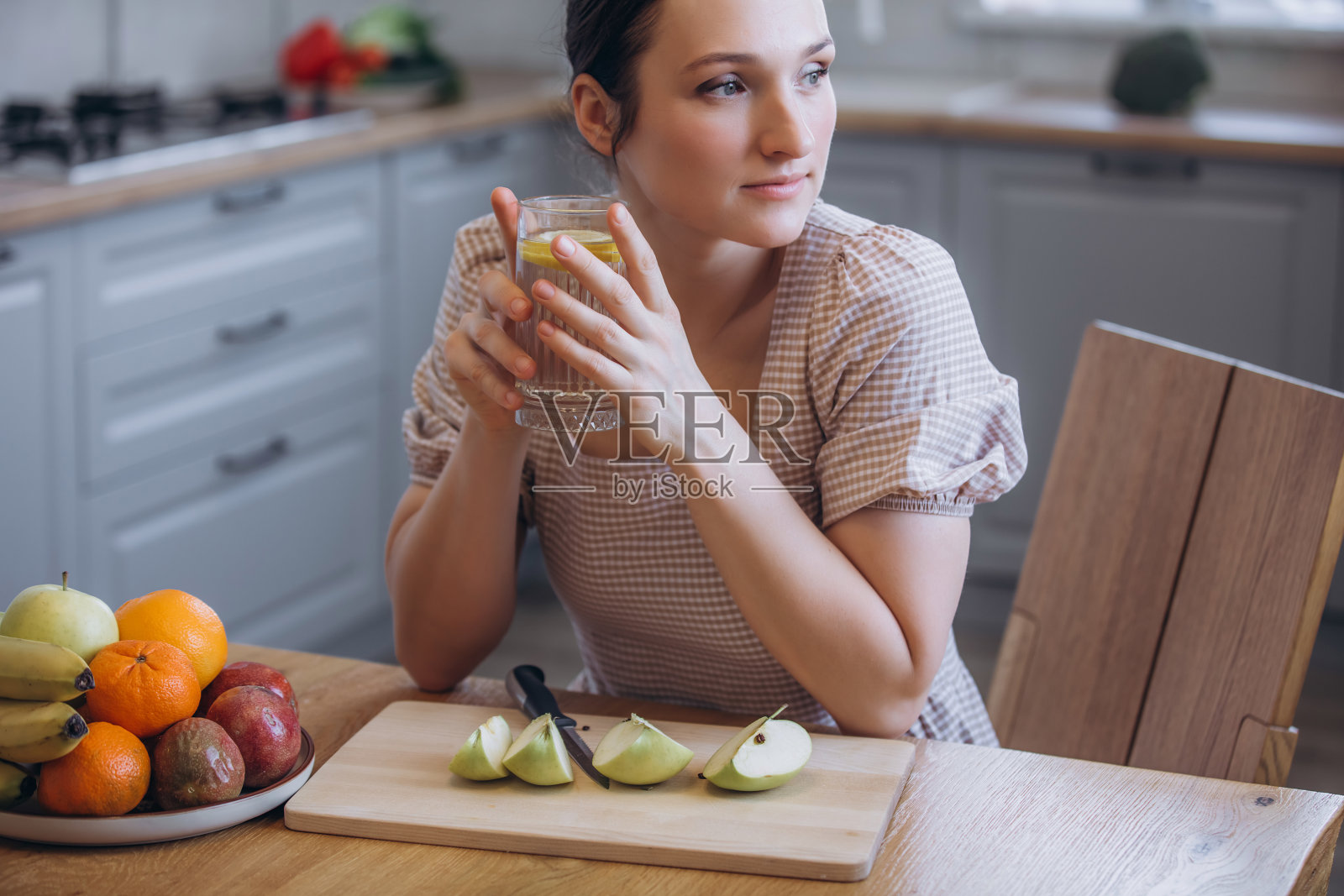 一位美丽的棕发女性在厨房里,手里拿着一杯加了柠檬和水果的水。照片摄影图片