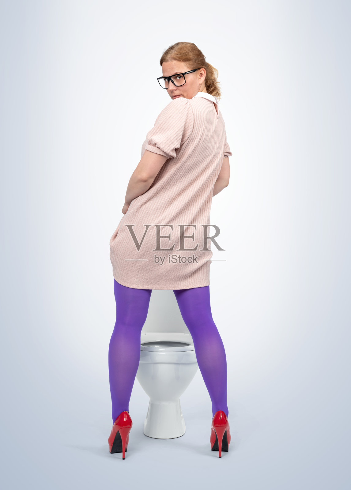 一位戴眼镜的女性穿着浅粉色连衣裙和大胆的紫色紧身裤,俏皮地站在厕所前。她穿着红色高跟鞋,自信地回头,背景是浅蓝色的。照片摄影图片