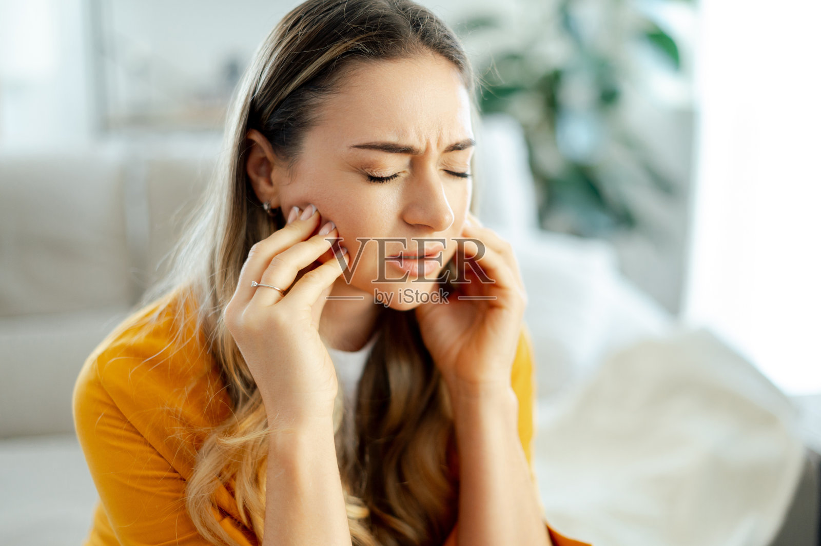 牙痛、牙科疼痛或下颌不适的概念。一个穿着黄色衬衫的年轻白人女性坐在客厅的沙发上,双手按着两颊,面露痛苦表情。照片摄影图片