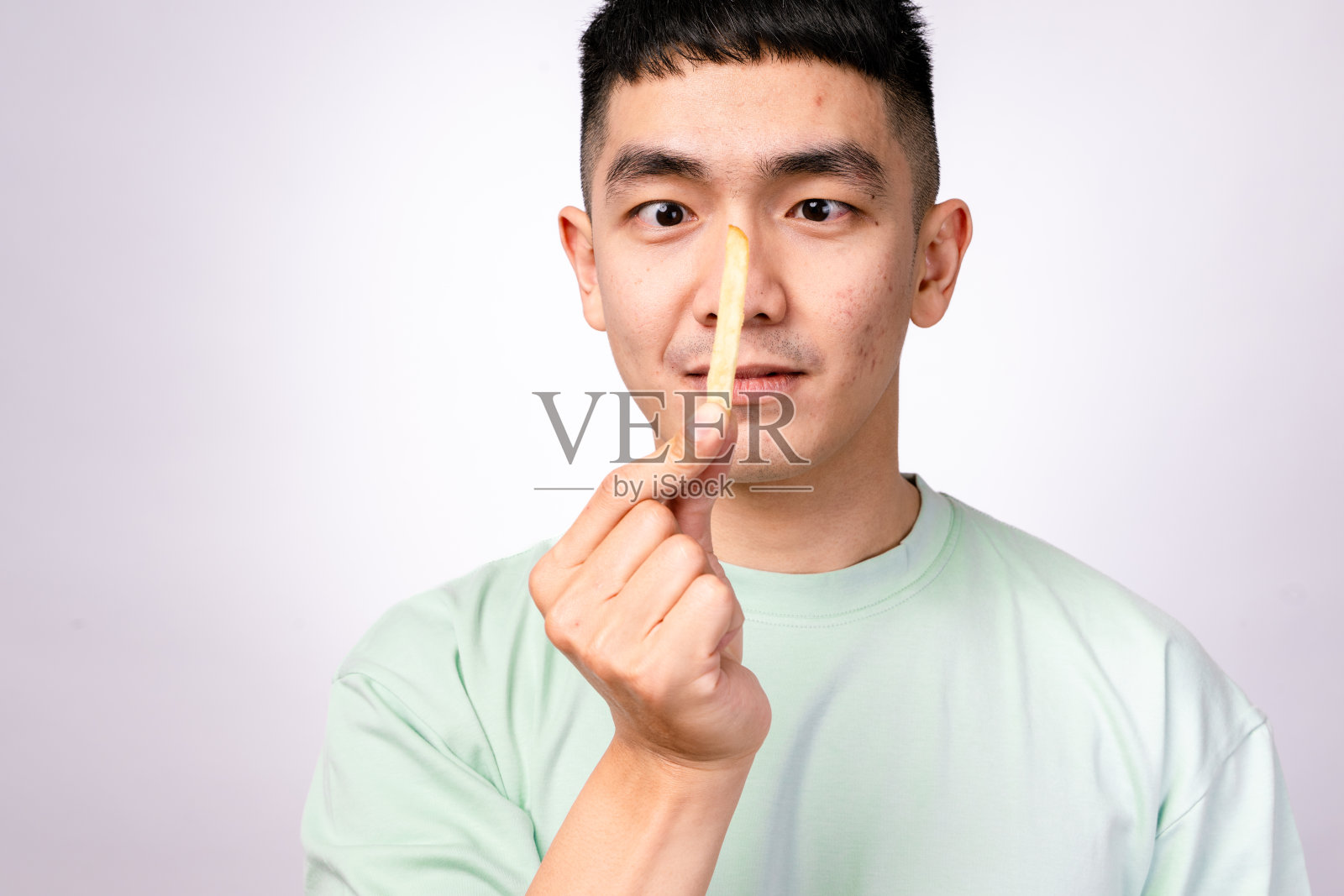 一位穿着浅绿色衬衫的亚洲男子正在吃薯条,同时手里拿着一个红色的薯条容器。他看起来很顽皮,侧目而视。背景是白色的,薯条看起来酥脆可口。照片摄影图片