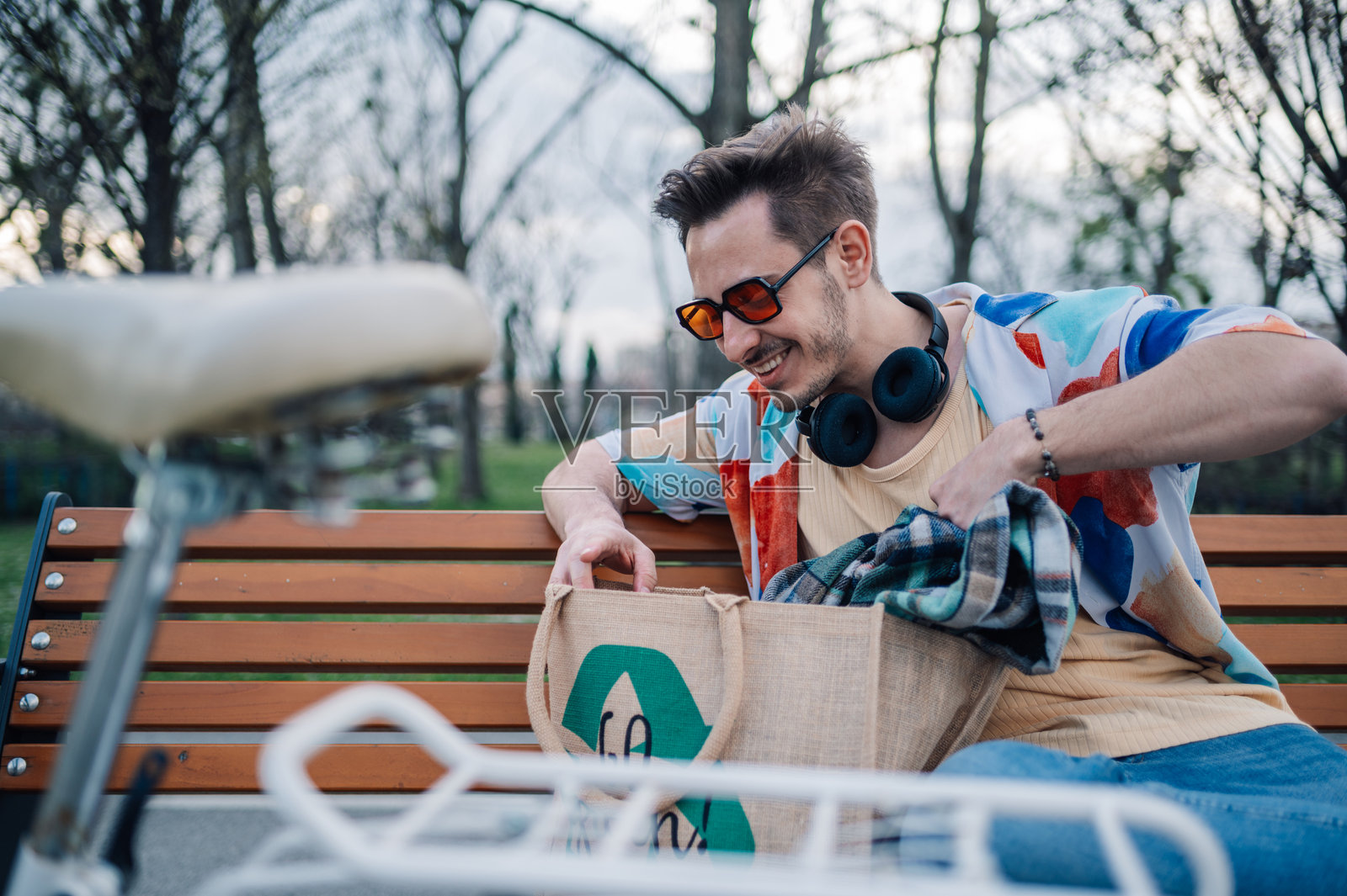 年轻男子坐在公园的长椅上,把衣物放进可重复使用的袋子里照片摄影图片