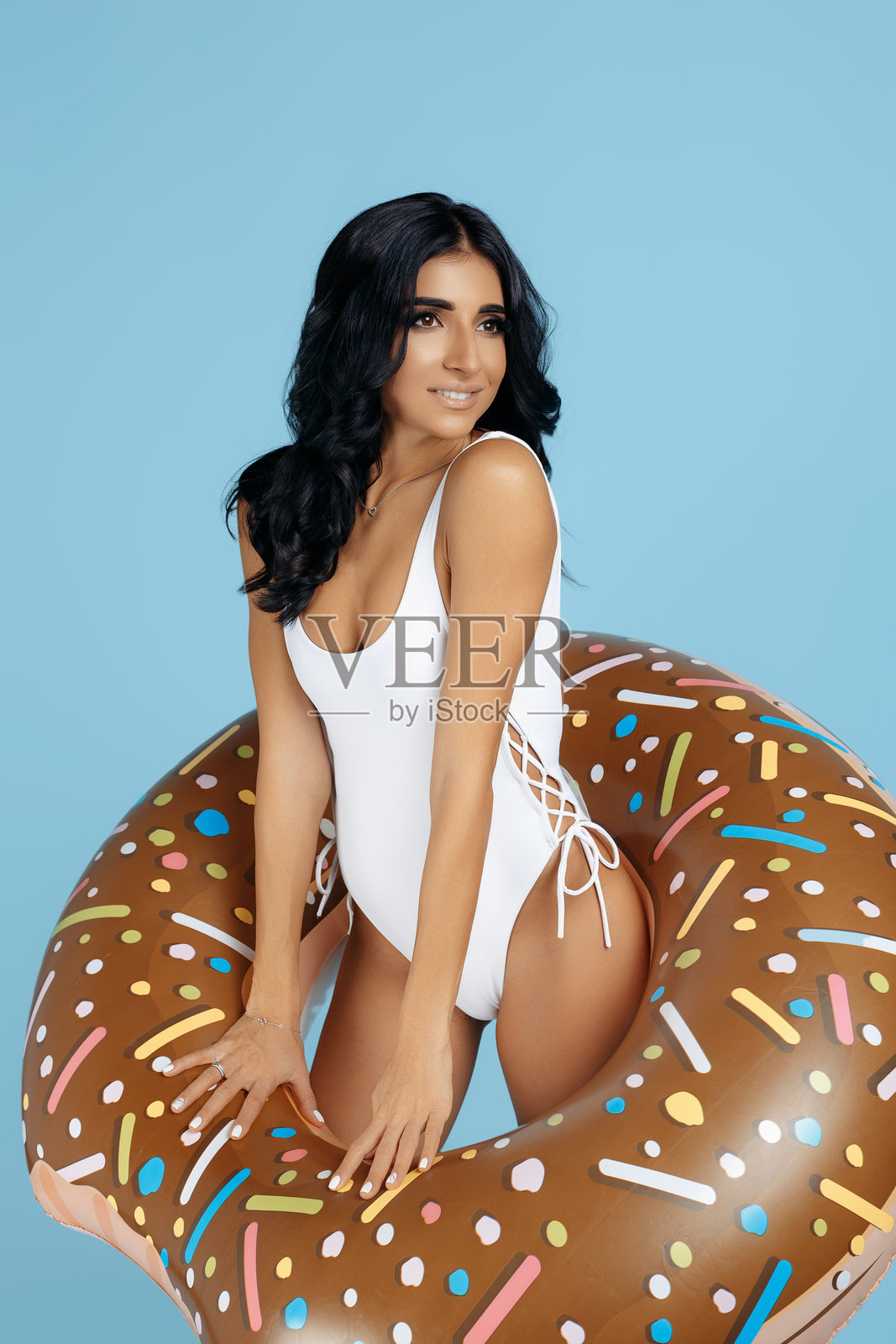 时尚的年轻女性穿着泳衣,开心地摆姿势,手持蓝色背景下的甜甜圈充气圈。照片摄影图片
