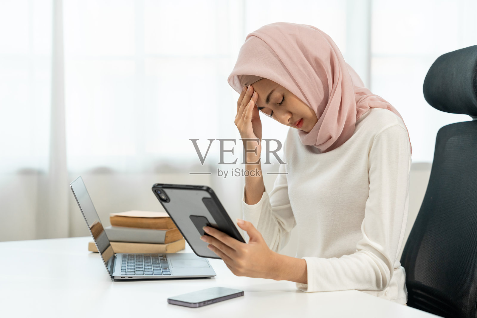 感到压力的亚洲穆斯林女商人手持平板电脑。伊斯兰女性在使用笔记本电脑时,盯着平板电脑屏幕感到压力。照片摄影图片
