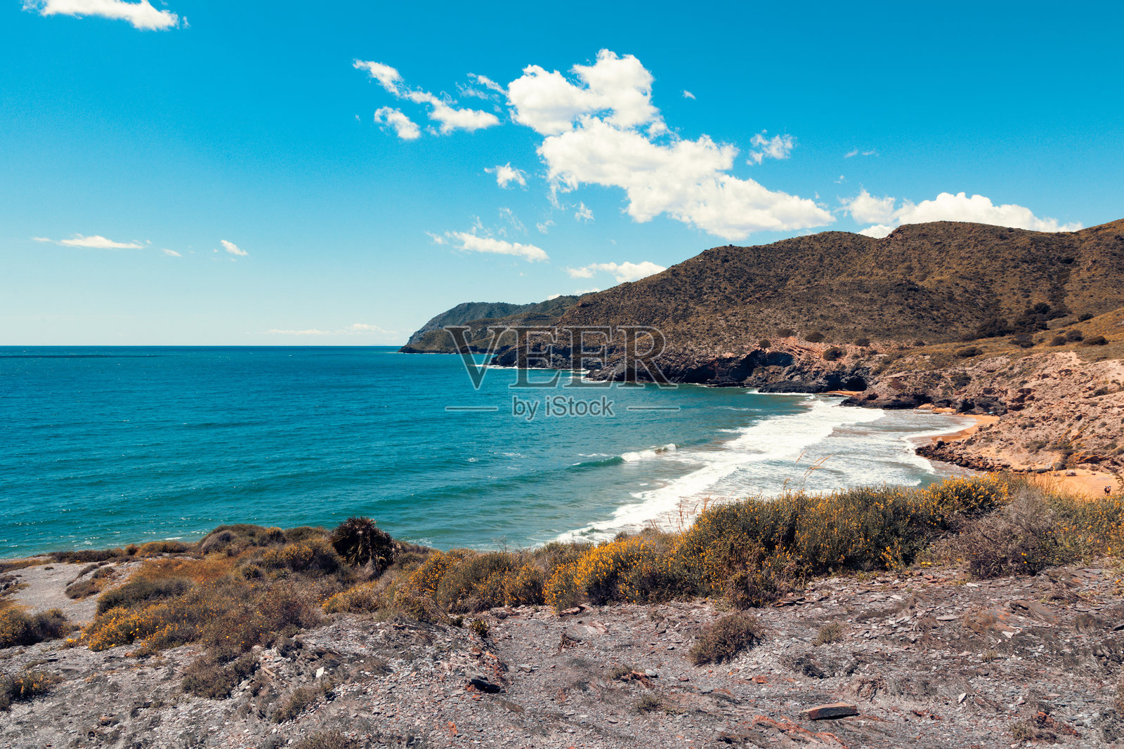 西班牙穆尔西亚的卡尔布兰克宁静海湾,金色沙滩、郁郁葱葱的海岸植被、碧蓝的海水和在明亮阳光下散布的云朵下的壮丽山丘。照片摄影图片