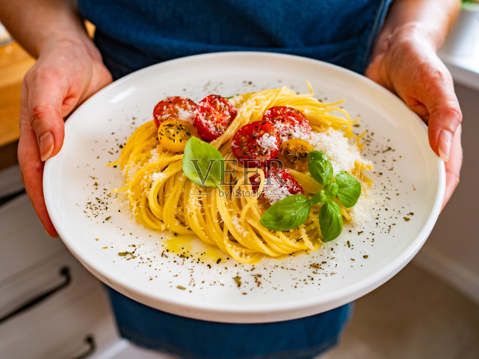 女人手持意大利面盘,上面撒有帕尔马干酪和新鲜樱桃番茄,摆放在木桌上。照片摄影图片
