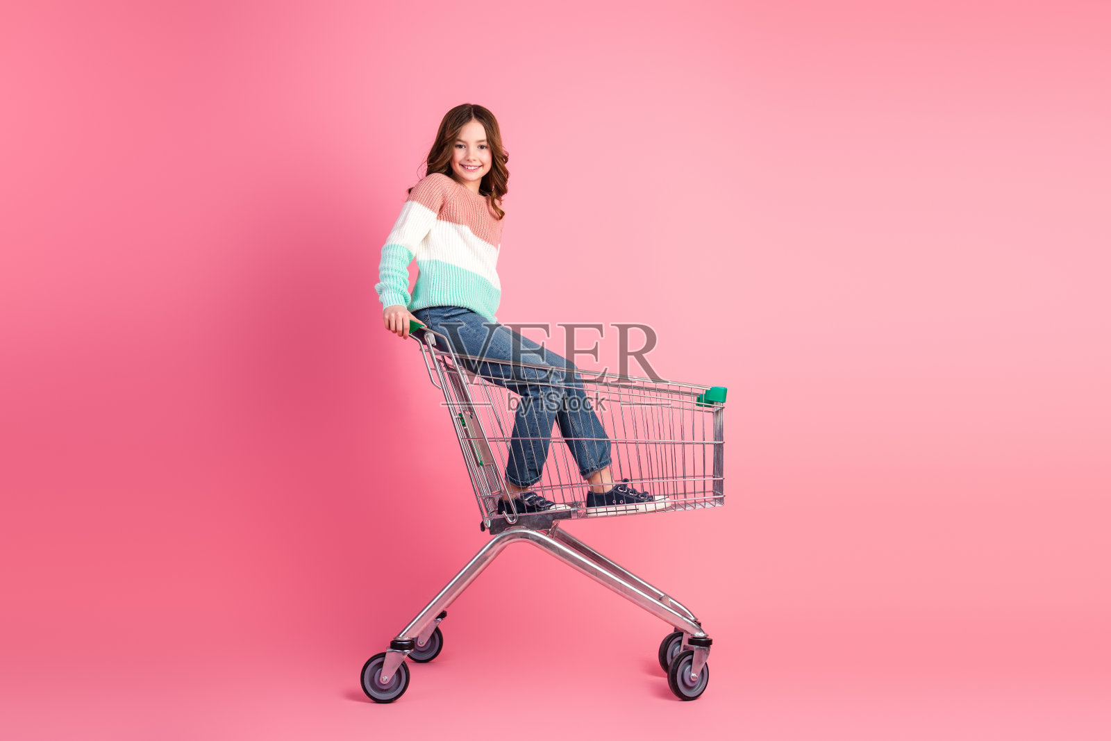 可爱的小女孩在粉色背景下顽皮地坐在购物车里,展现青春、休闲和时尚风格。照片摄影图片