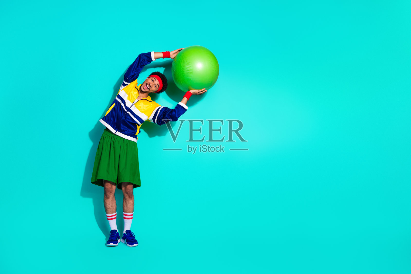 年轻男子穿着色彩鲜艳的复古运动服,热情地在充满活力的青绿色工作室背景下使用健身球锻炼。照片摄影图片