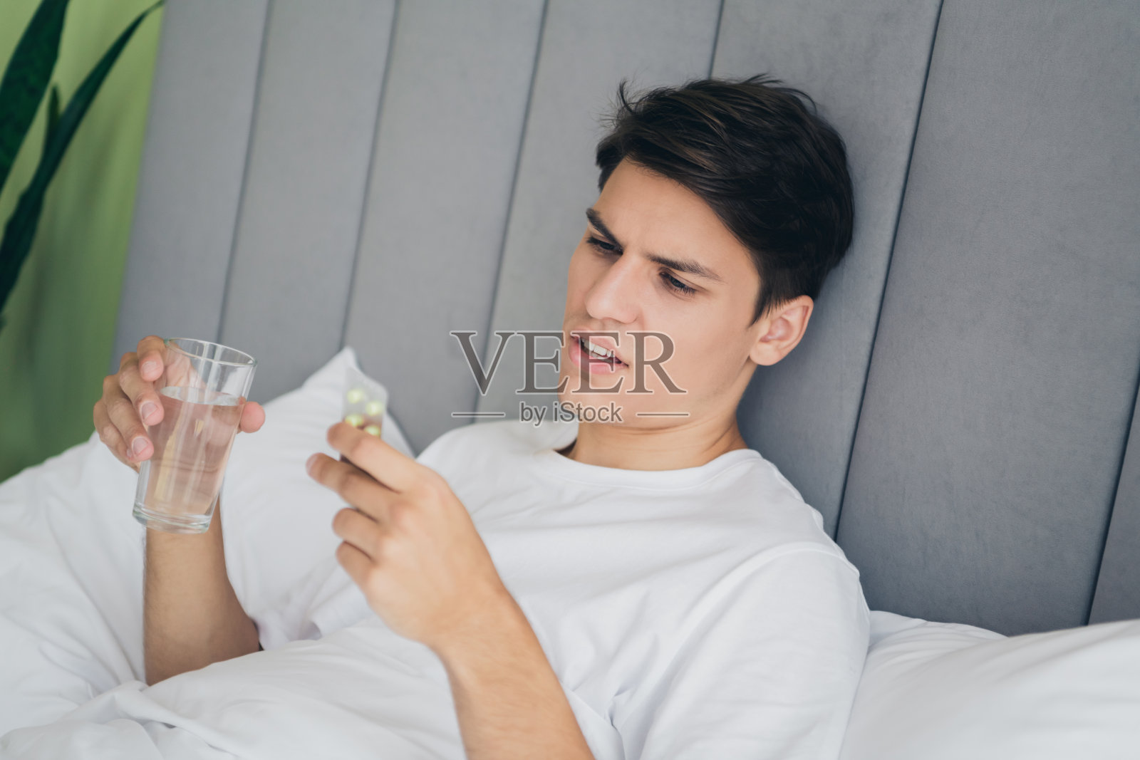 年轻男子坐在床上,手里拿着一杯水,仔细查看药物条,神情沉思。照片摄影图片