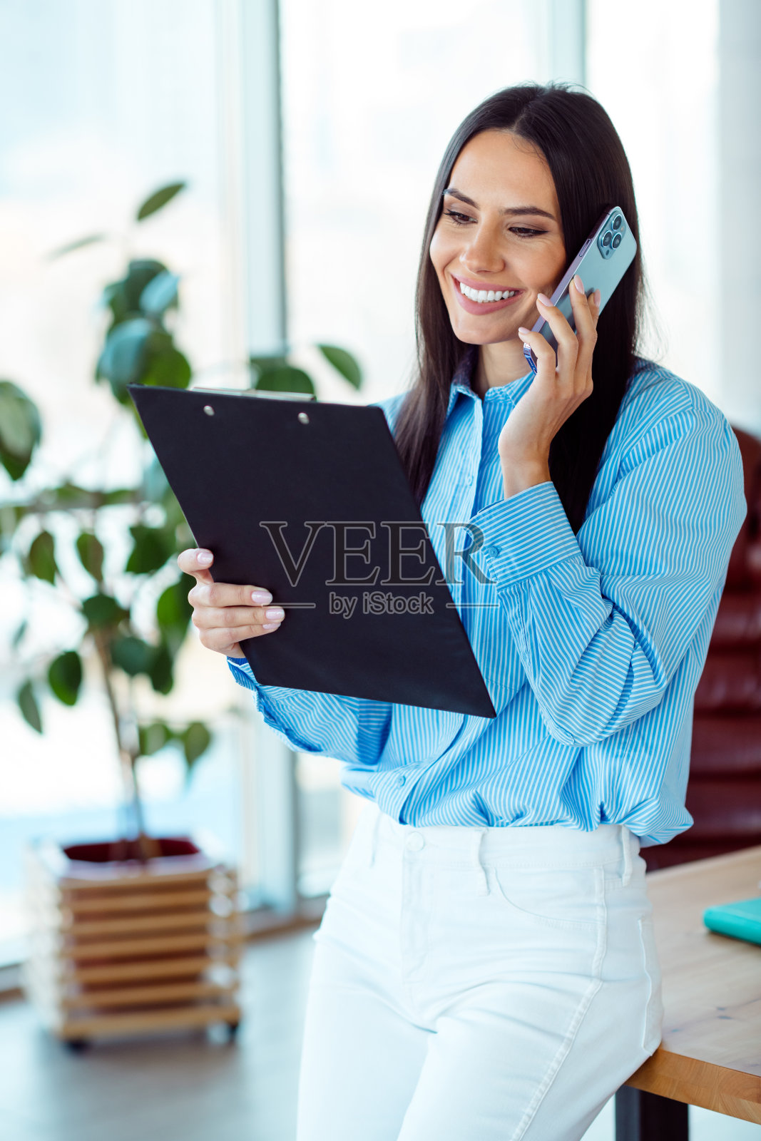 自信的年轻女商人在现代办公室工作,穿着休闲职业装,通过电话讨论工作并审阅文件。照片摄影图片