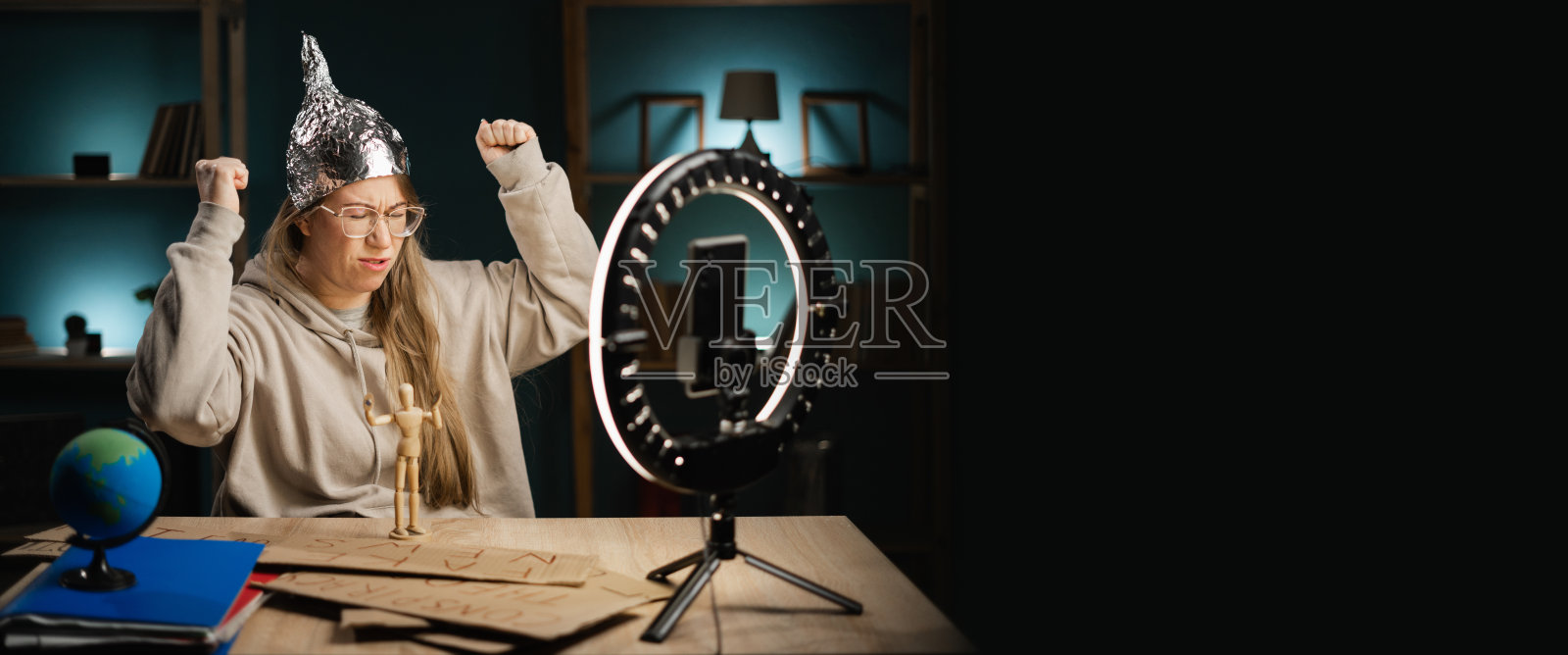 戴眼镜的疯狂阴谋论博主在家中使用智能手机和环形灯进行直播,双拳紧握,像个胜利者。横幅。照片摄影图片