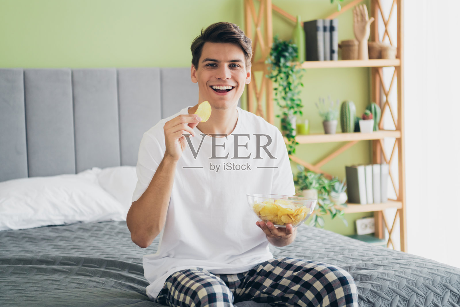 年轻男子在温馨的卧室里享受悠闲时光,坐在床上,沐浴在愉快的晨光中,穿着休闲睡衣。照片摄影图片