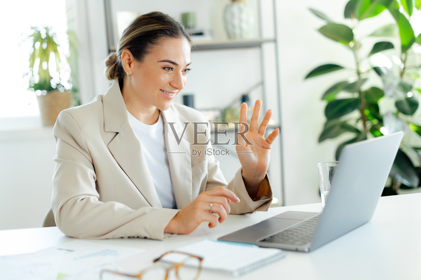 自信成功的白人女商人穿着时尚的西装,微笑着在办公室的工作桌前坐着,通过笔记本电脑与同事或客户进行视频通话,挥手致意。照片摄影图片