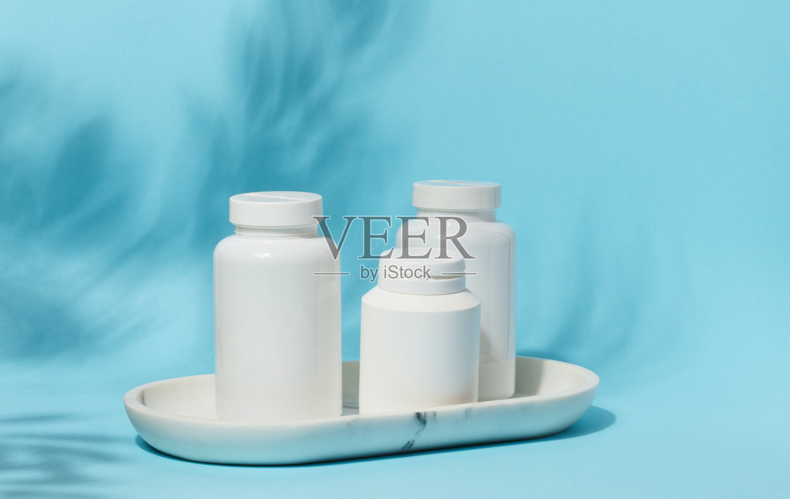 三个不同大小的白色塑料药瓶放在白色大理石托盘上,背景是淡蓝色,带有细微的叶子阴影。照片摄影图片