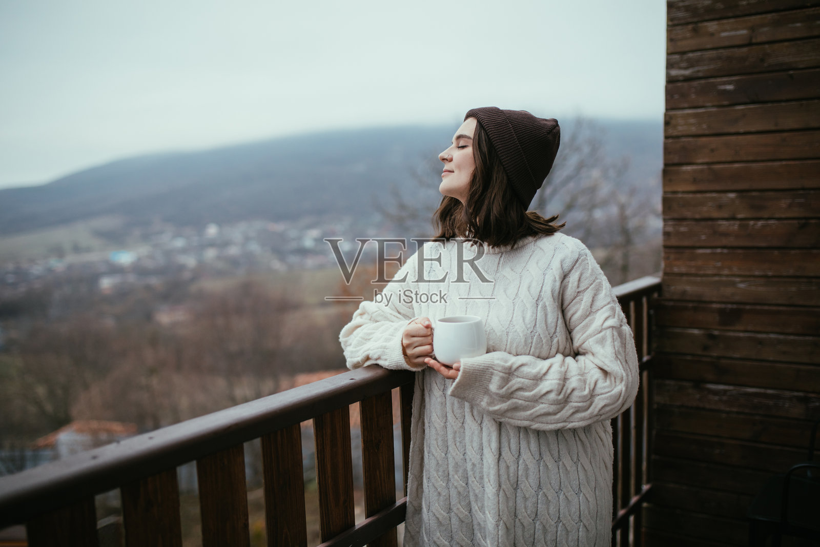 年轻女性穿着白色针织毛衣,手握白色茶杯,站在木屋的阳台上,俯瞰山景。照片摄影图片