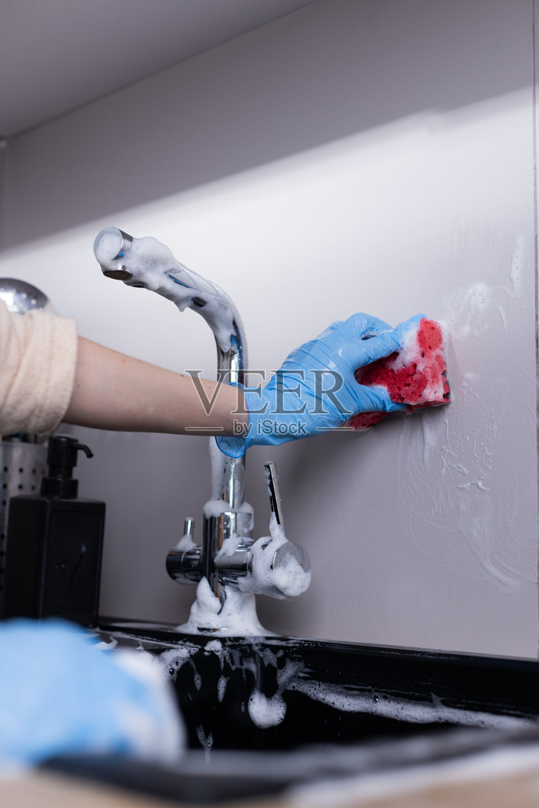 一只戴着蓝色手套的手在明亮的厨房环境中用粉色海绵和肥皂清洁靠近水槽的墙壁照片摄影图片