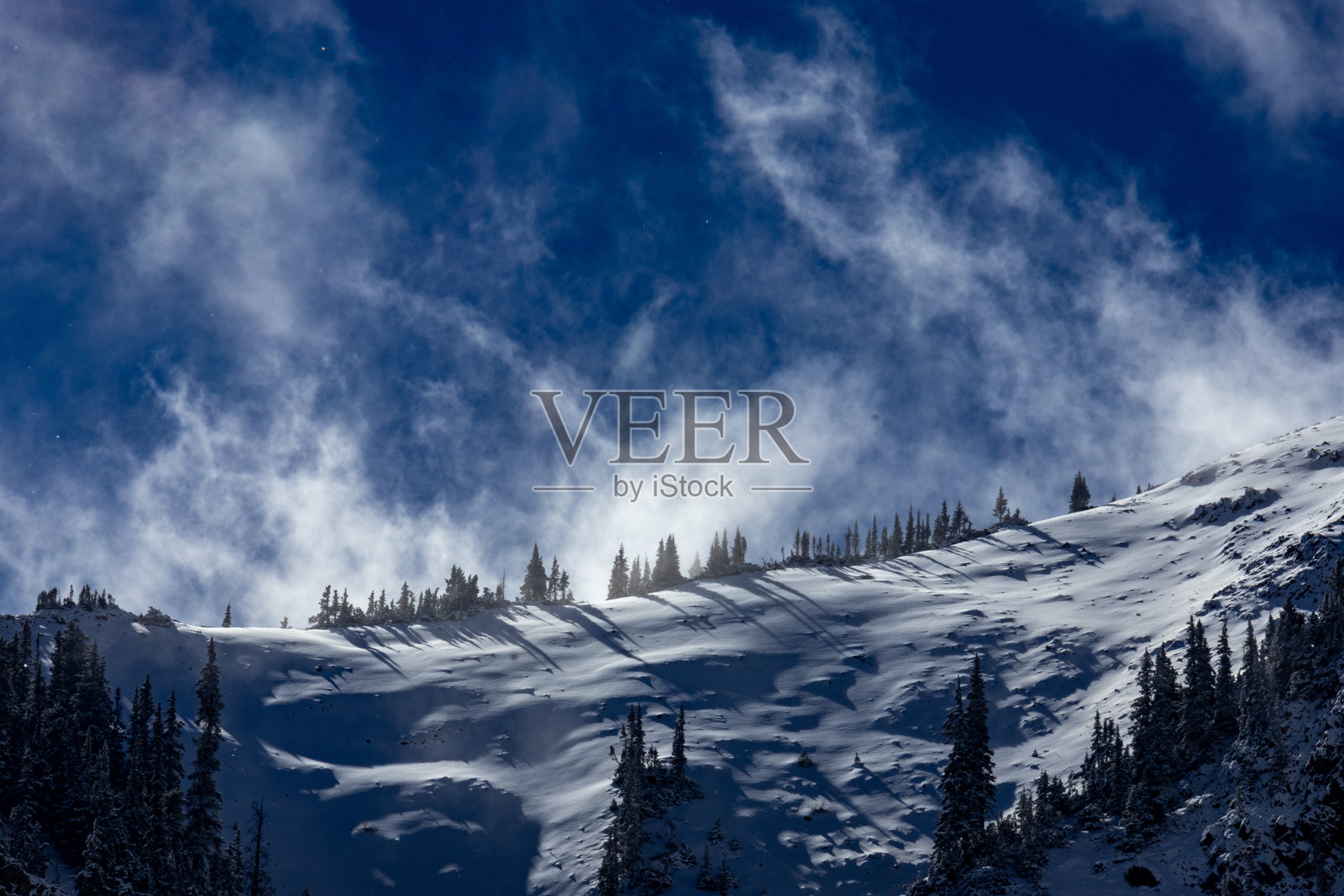 在明亮的蓝天下,雪山景观与漂浮的云朵交织,映衬着傍晚的光线。照片摄影图片