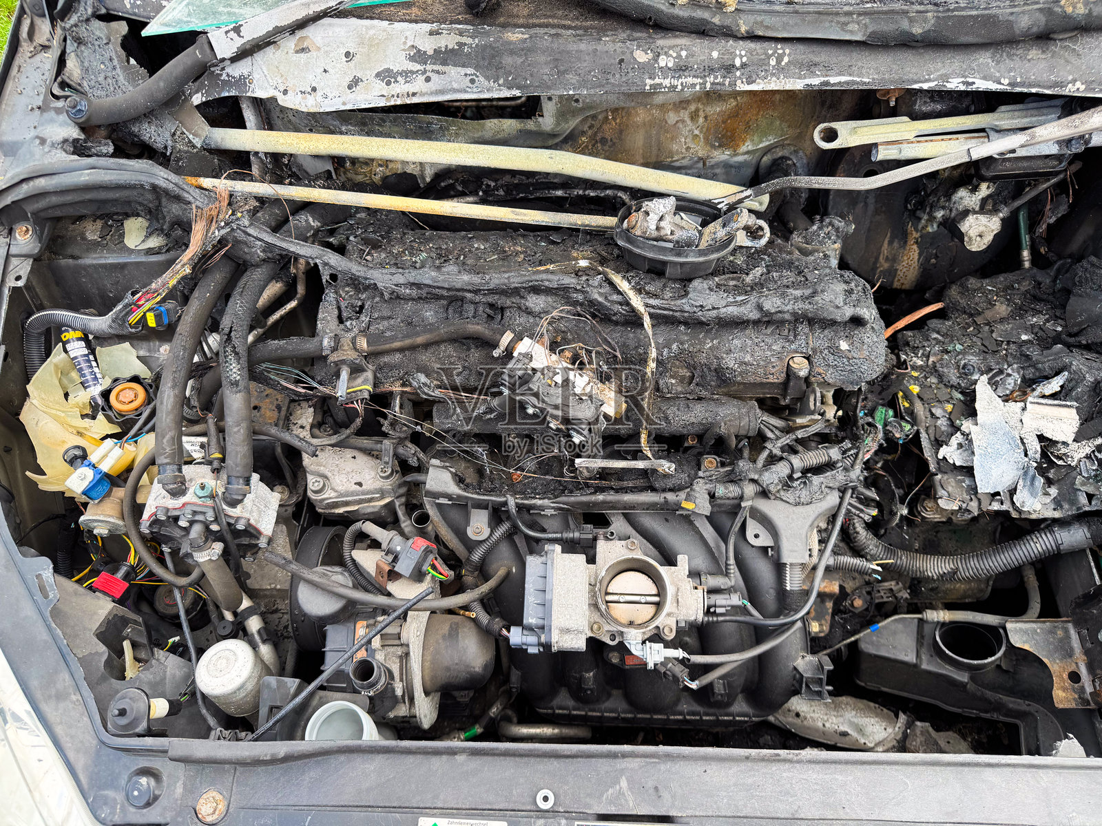 烧毁的汽车发动机舱,显示出火灾损坏的发动机块、融化的电线、烧焦的软管和严重热损伤的暴露进气歧管照片摄影图片