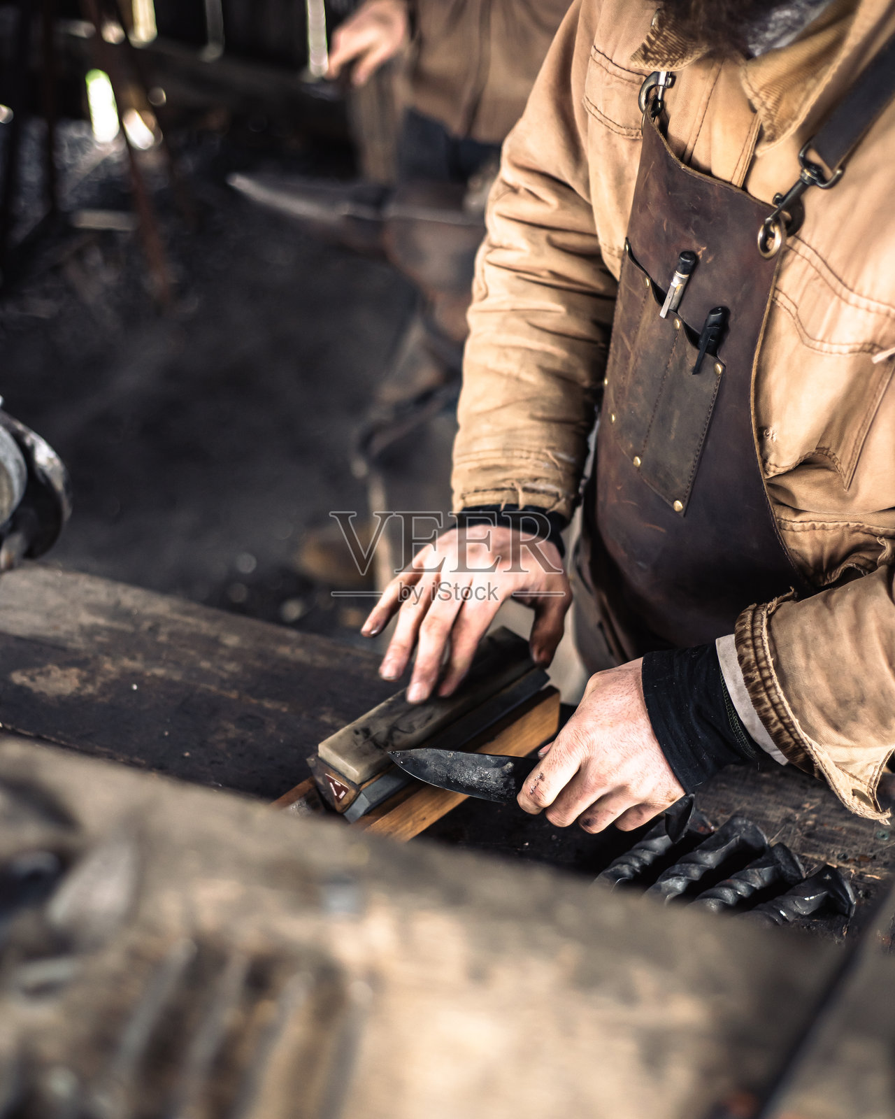 密苏里州曼斯菲尔德的传统工坊里,身穿皮围裙的铁匠正在磨刀,白人工匠在磨石上磨刀刃,打磨钢工具的边缘。照片摄影图片
