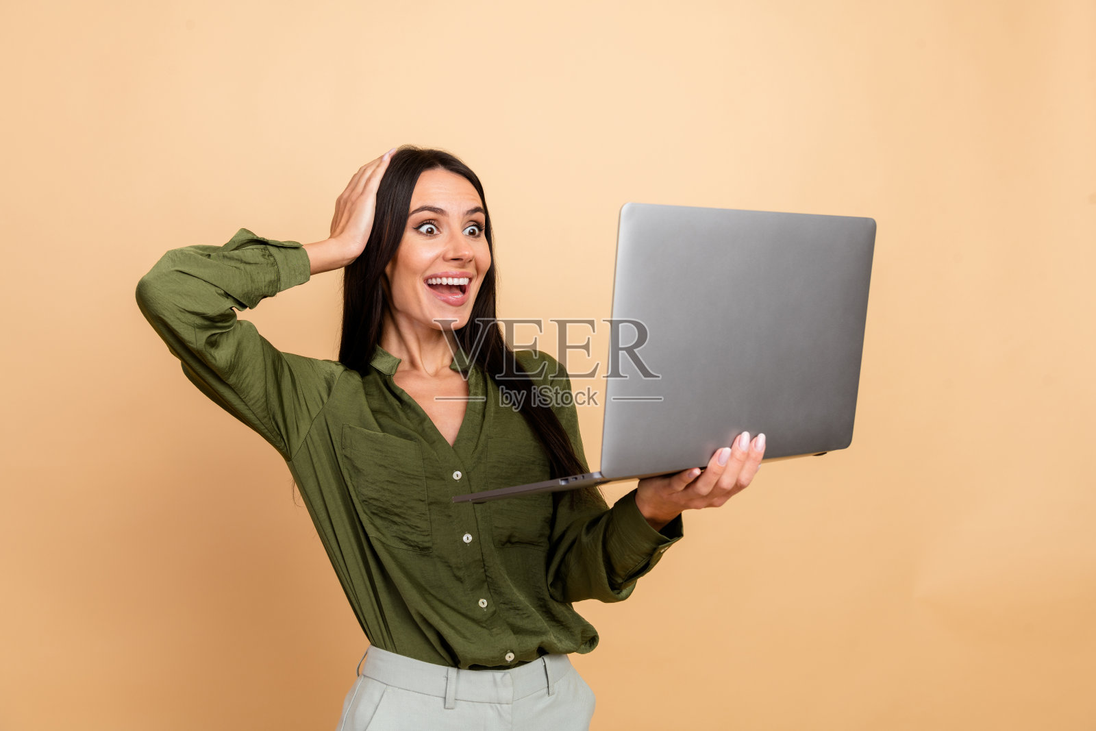 兴奋的年轻女商人穿着绿色衬衫,手持笔记本电脑,背景为米色,表现出惊讶和快乐的手势。照片摄影图片