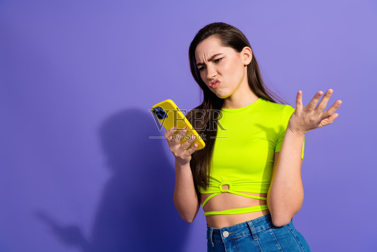 穿着鲜艳服装、面露困惑表情的年轻女性在明亮的紫色背景前使用智能手机,展现出困惑的状态照片摄影图片