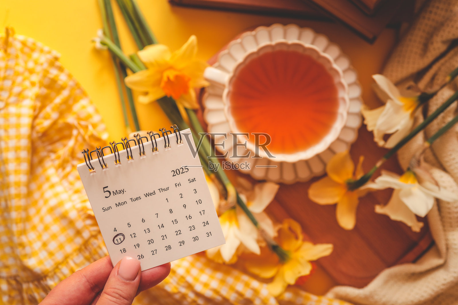 手握着一个小型的2025年5月日历,11号被圈出,旁边是一杯茶、黄水仙花和舒适的织物,营造出温暖的春季场景照片摄影图片