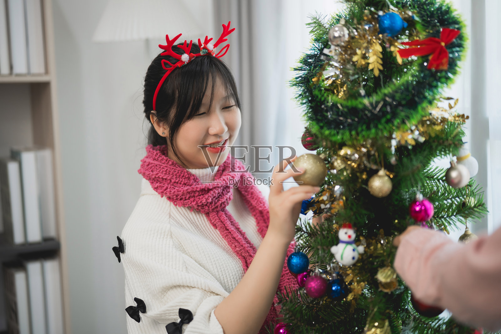 快乐的亚洲年轻女性在舒适的客厅里装饰圣诞树,挂上装饰品和灯光,庆祝节日的欢乐气氛。照片摄影图片