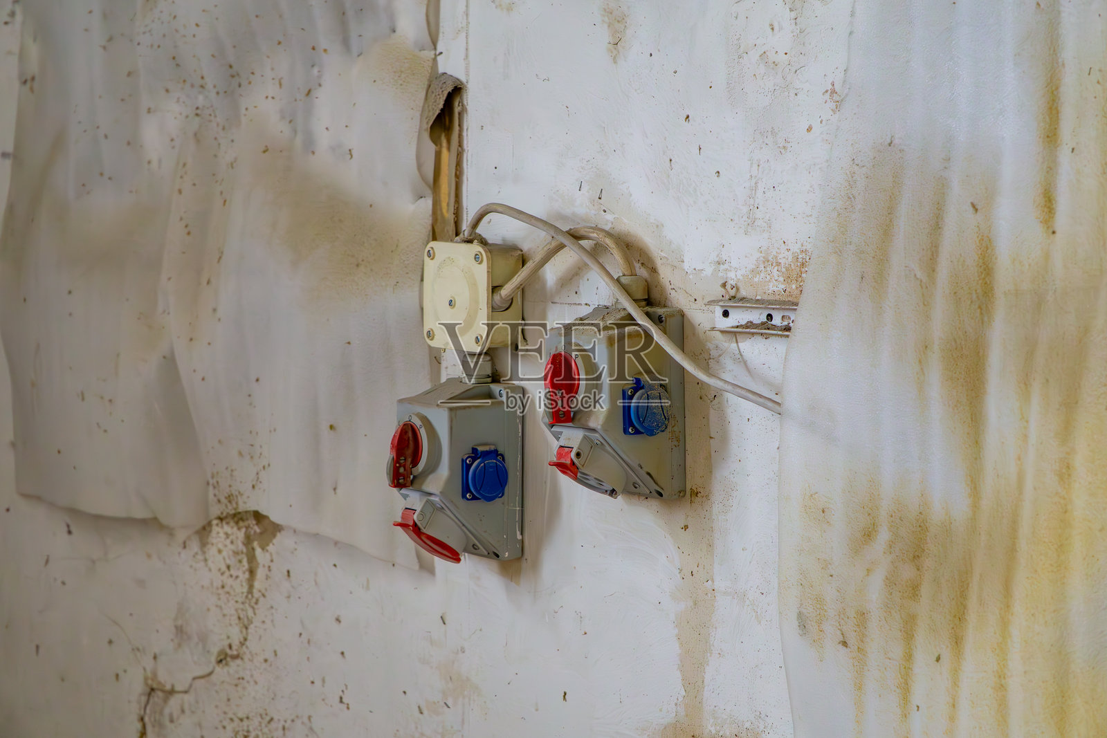 一面旧而肮脏的墙上安装着工业插座(电源插座)和电缆。插座有红色和蓝色,典型用于工业设施中的三相和单相安装。照片摄影图片