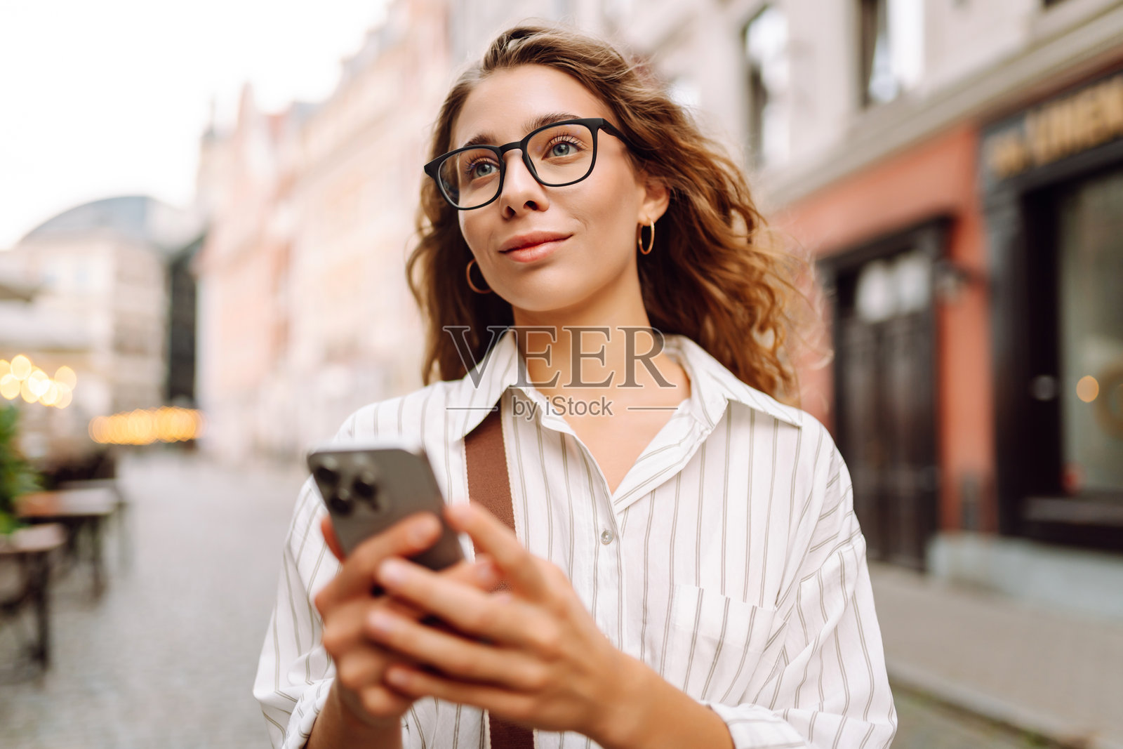 一位年轻女性穿着时尚的衣服,手握手机,走在阳光明媚的城市街道上。照片摄影图片