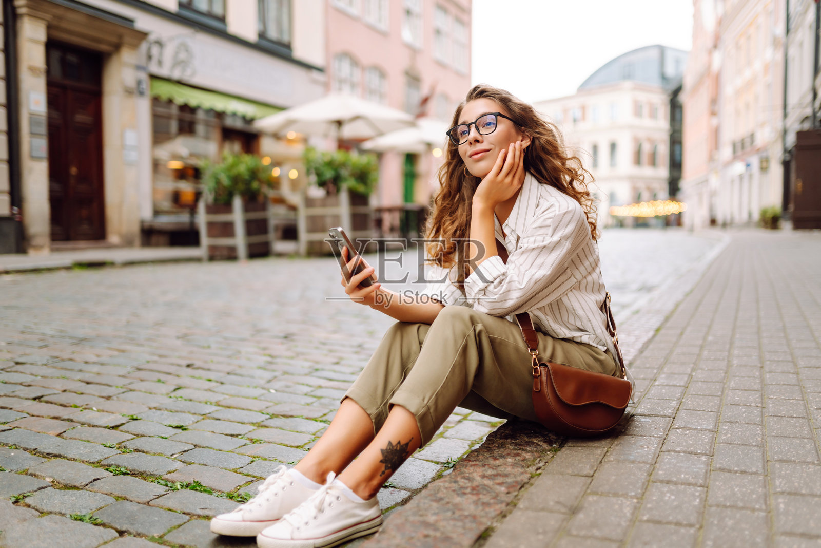 一位年轻女性穿着时尚的衣服,手中拿着手机,走在阳光明媚的城市街道上。照片摄影图片