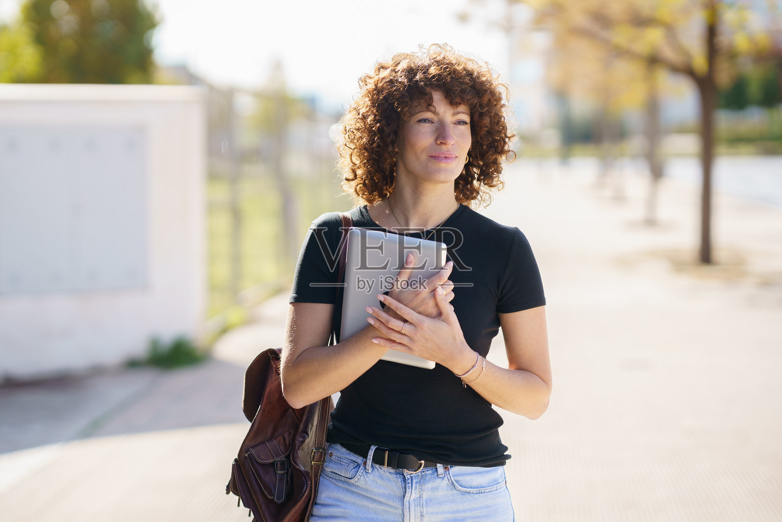 一位年轻女性,拥有美丽的卷发,走在户外,手里提着笔记本电脑和笔记本。照片摄影图片