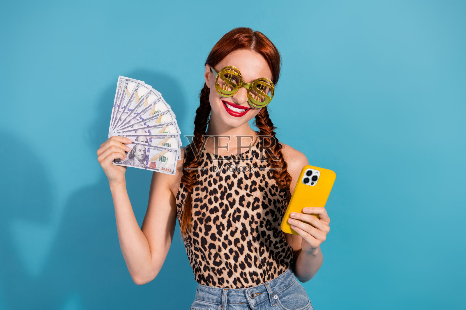 穿着豹纹上衣的年轻女性在蓝色背景前手持现金和手机,展示轻松愉快的夏日生活方式照片摄影图片
