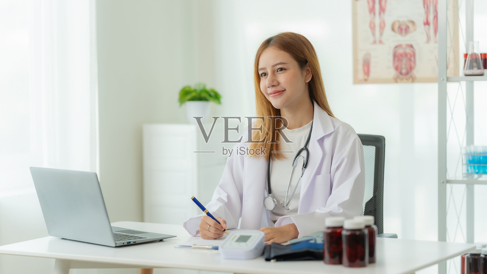 微笑的亚洲女性医生穿着白大褂和听诊器,坐在医生办公室的在线咨询桌旁照片摄影图片