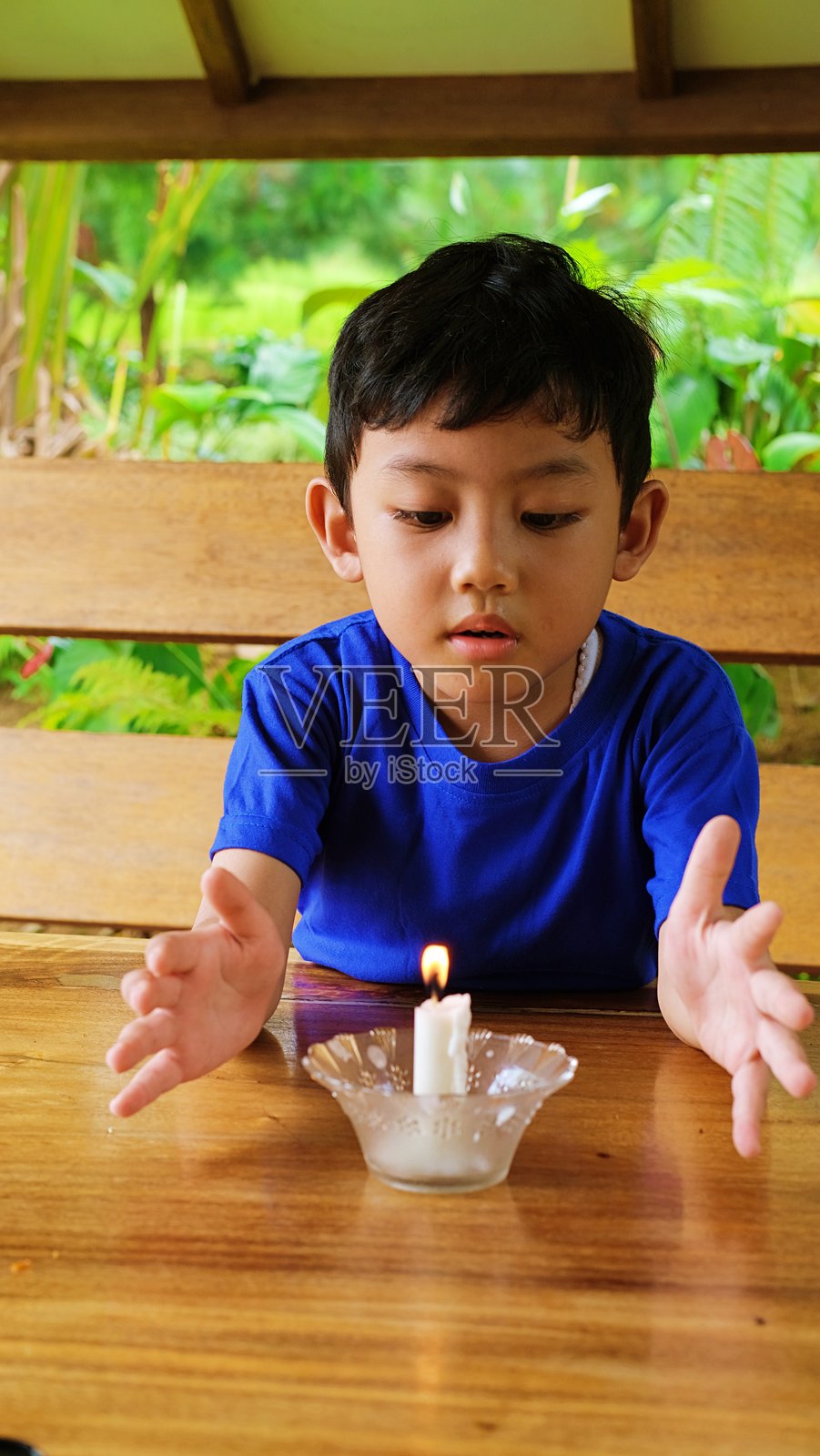 一个穿着蓝色衬衫的小男孩坐在户外,好奇地观察着木桌上的小蜡烛火焰,周围环绕着郁郁葱葱的绿树。照片摄影图片
