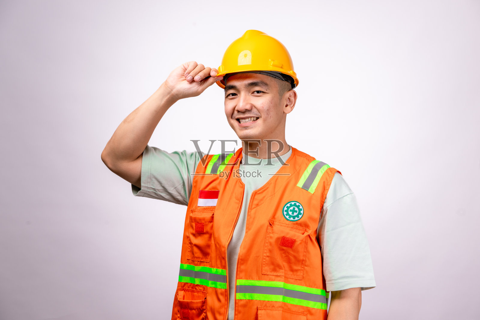 一名亚洲男性建筑工人戴着黄色安全帽和橙色安全背心,自信地双手叉腰,直视镜头。背景为白色。照片摄影图片