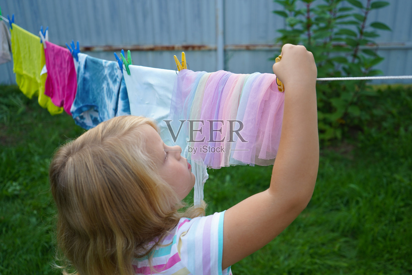 一个大约六岁的小女孩踮起脚尖,小心翼翼地把衣物夹在阳光明媚的下午衣绳上。照片摄影图片