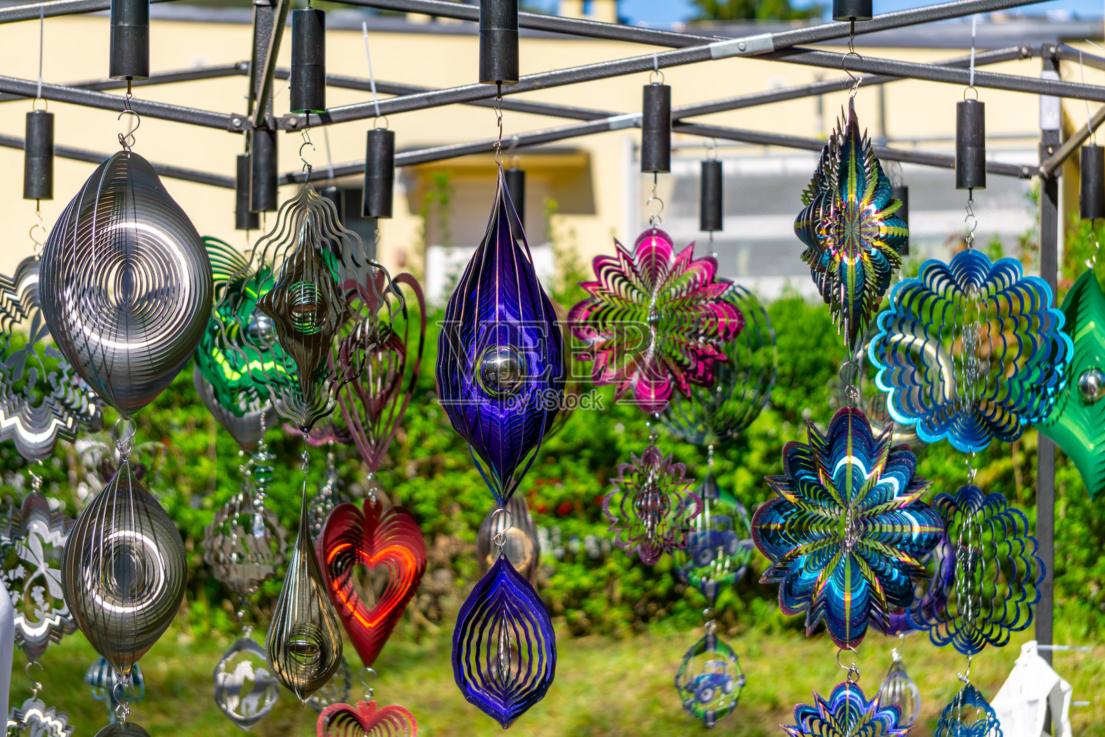 色彩斑斓的金属花园装饰,呈吊坠和风车的形状,能够反射光线。它们悬挂在一个框架上,背景是绿色的,适合在集市或户外市集时使用。照片摄影图片