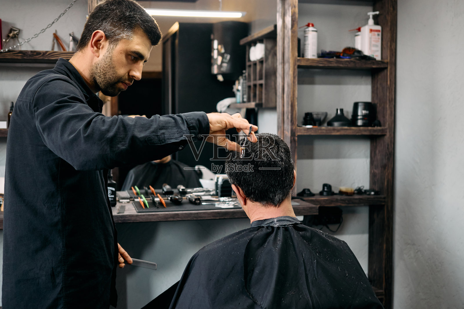 一位技艺高超的理发师在现代理发店中专心为顾客剪发。气氛热闹,工具和产品整齐地摆放在附近,展现出专业的环境。照片摄影图片
