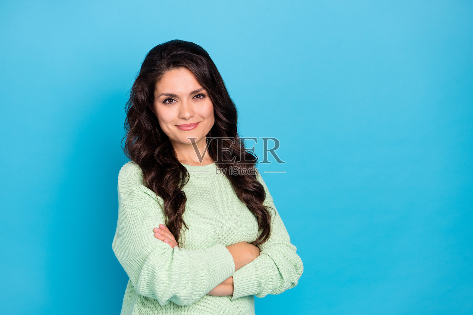 迷人、愉快的波浪卷发女性穿着温暖的毛衣,双臂交叉,背景为明亮的蓝色。照片摄影图片