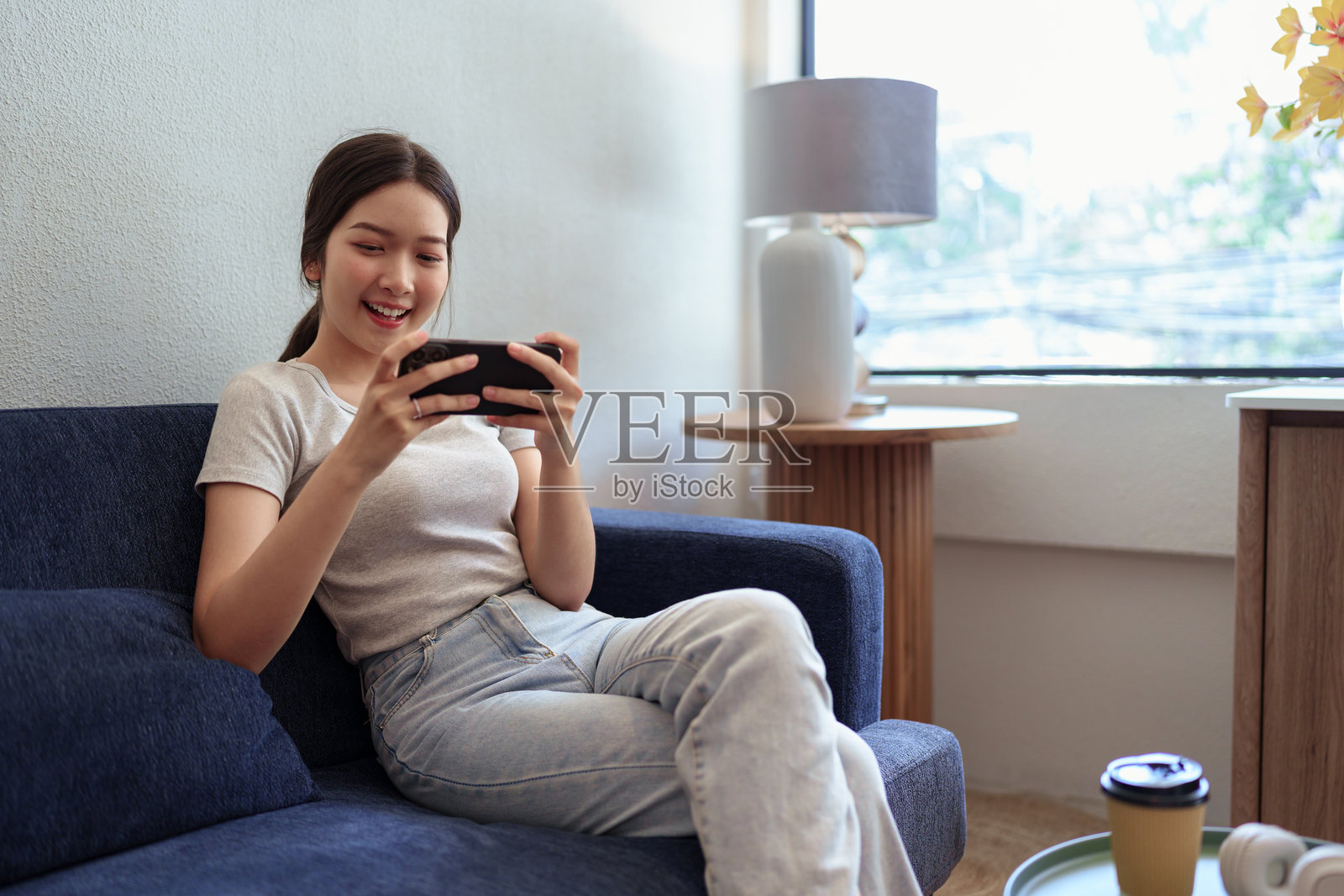 年轻女性在舒适的阳光咖啡角落里坐在沙发上享受手机游戏。照片摄影图片