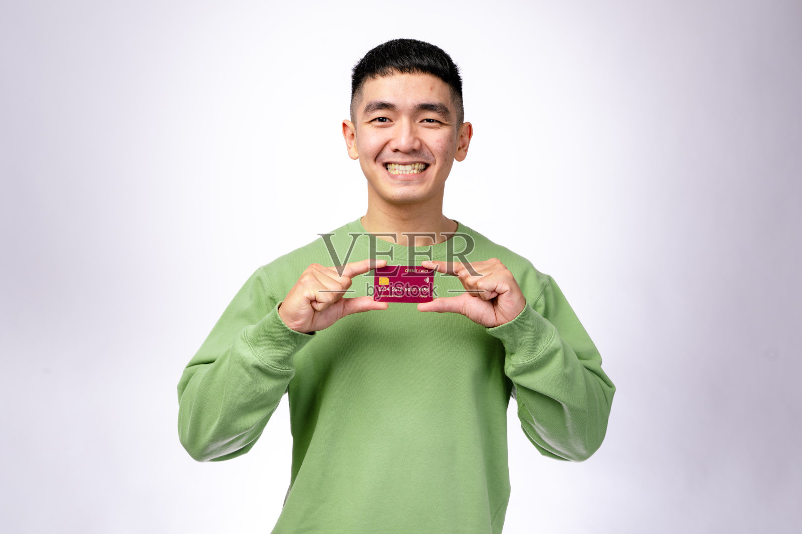 一位微笑的亚洲男子穿着绿色卫衣,手持一张红色信用卡靠近脸部,显得自信。他站在简单的白色背景前,象征着银行业务和无现金交易。照片摄影图片