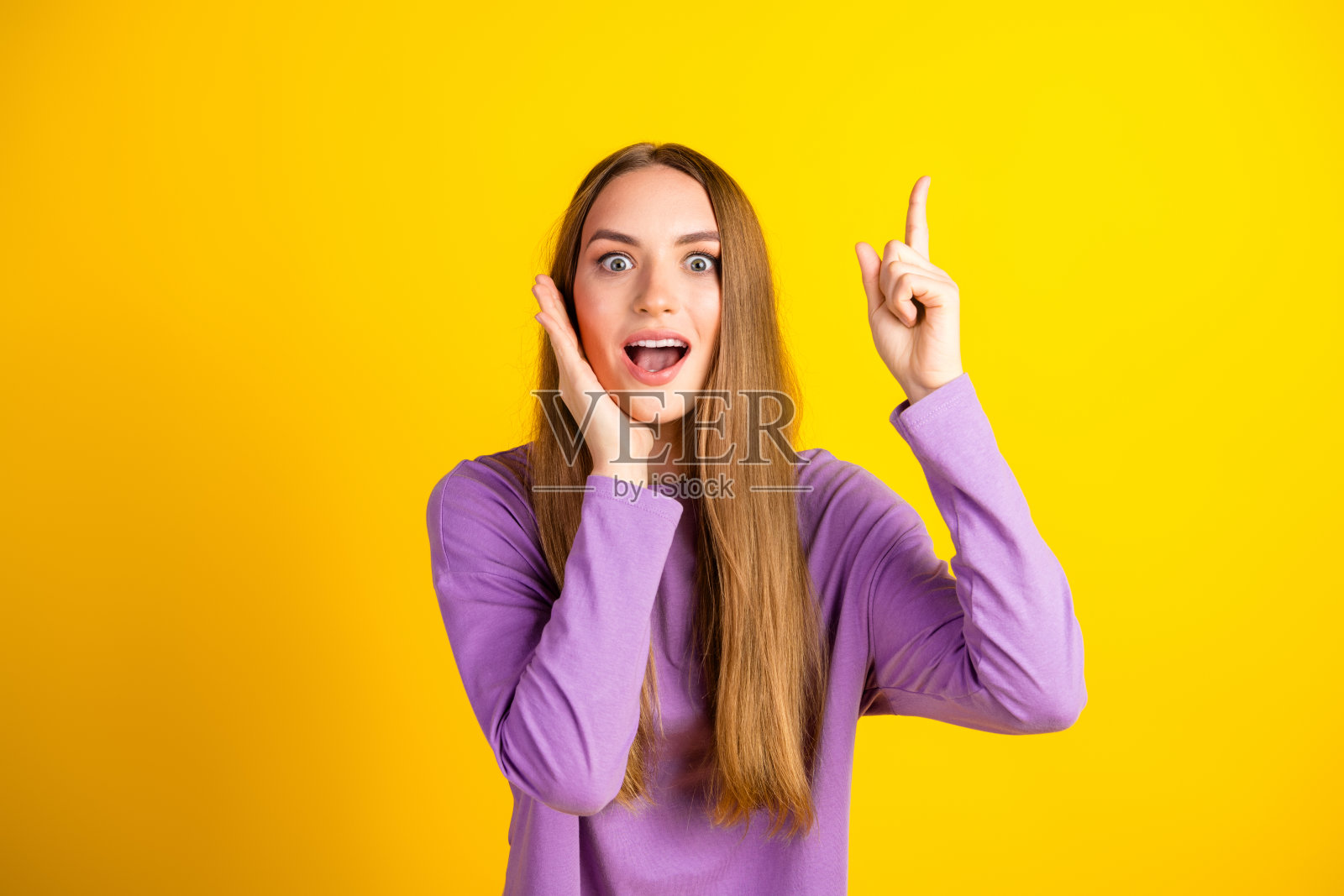 兴奋的年轻女性穿着休闲紫色服装,热情地指向上方,背景是鲜艳的黄色。照片摄影图片