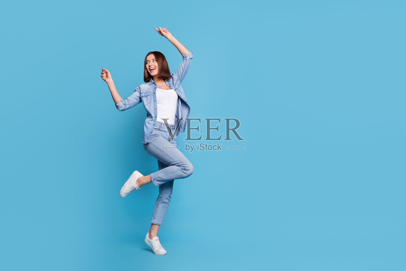 甜美闪亮的女士穿着牛仔衬衫,在蓝色背景下的空旷空间中跳舞照片摄影图片
