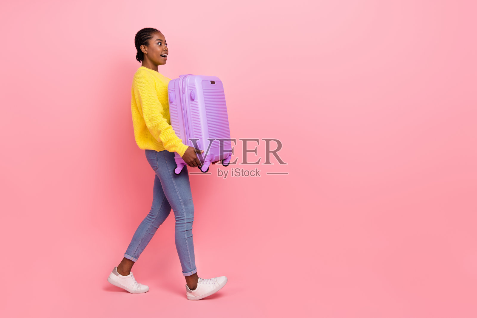 兴奋的疯狂女孩全身侧面肖像,手持大行李箱,望向空旷的空间,背景为粉色。照片摄影图片