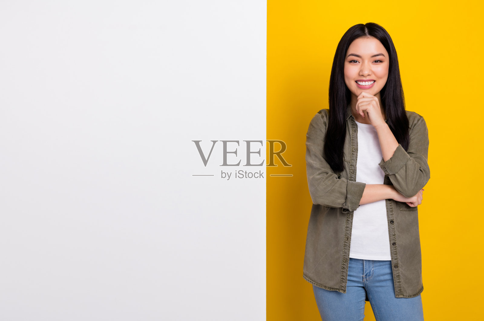 可爱的年轻棕发女孩穿着卡其色衬衫,站在大电话旁,背景为黄色。照片摄影图片