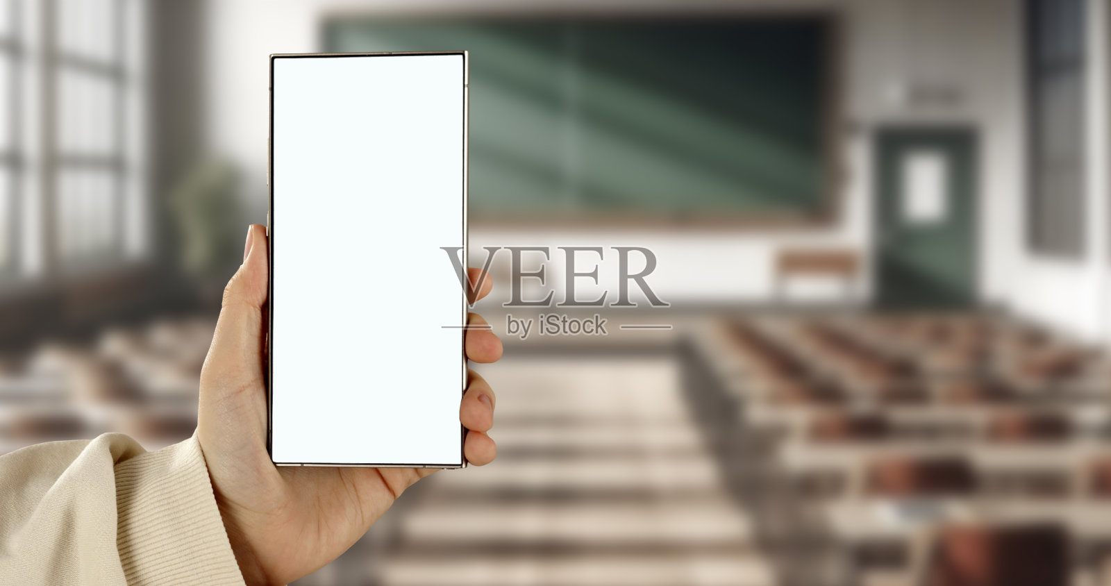 一只手握着一部屏幕空白的智能手机,背景是排成行的空桌子教室。照片摄影图片