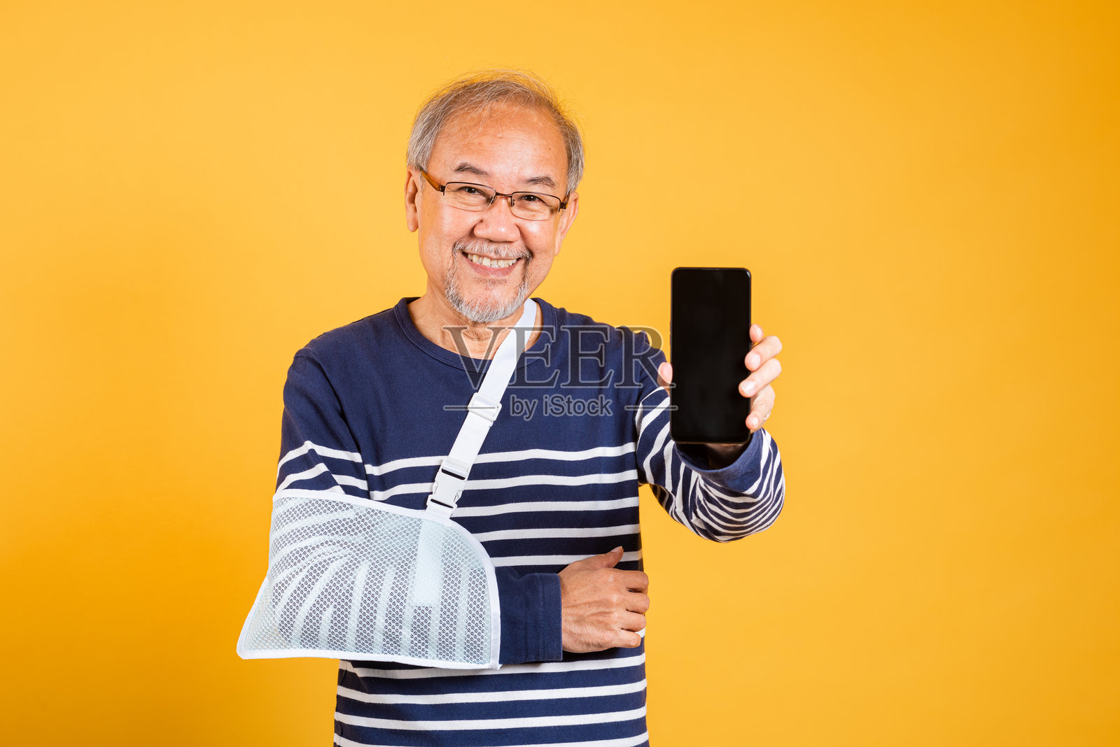 亚洲老年男子在黄色背景下展示空白手机屏幕,手臂打着夹板,正在接受治疗。照片摄影图片