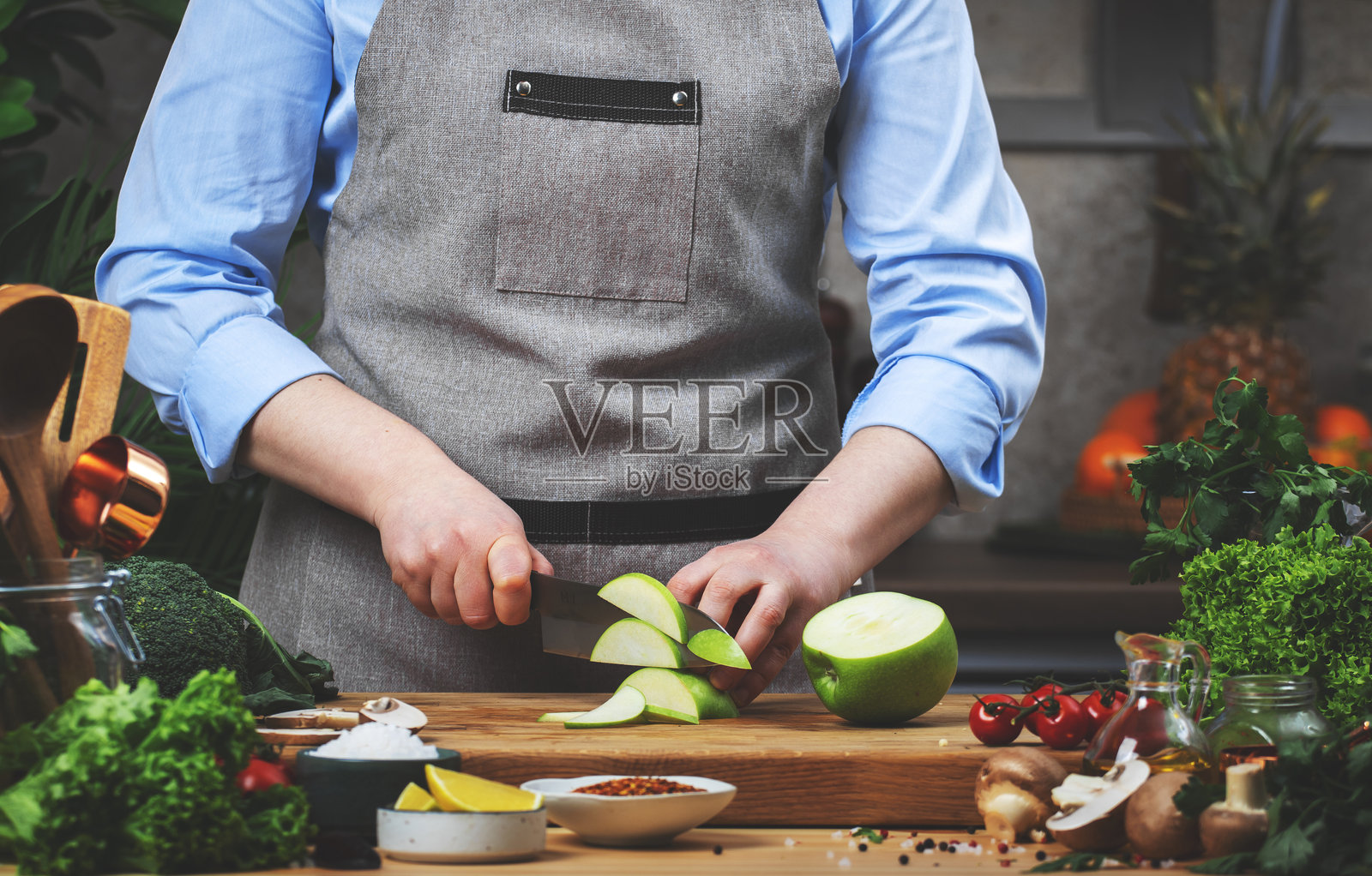 在厨房烹饪食物。厨师在木桌上将绿色苹果切成片,周围有厨房用具、蔬菜、香草和其他食材。健康的素食饮食,烹饪,食谱,博客。照片摄影图片