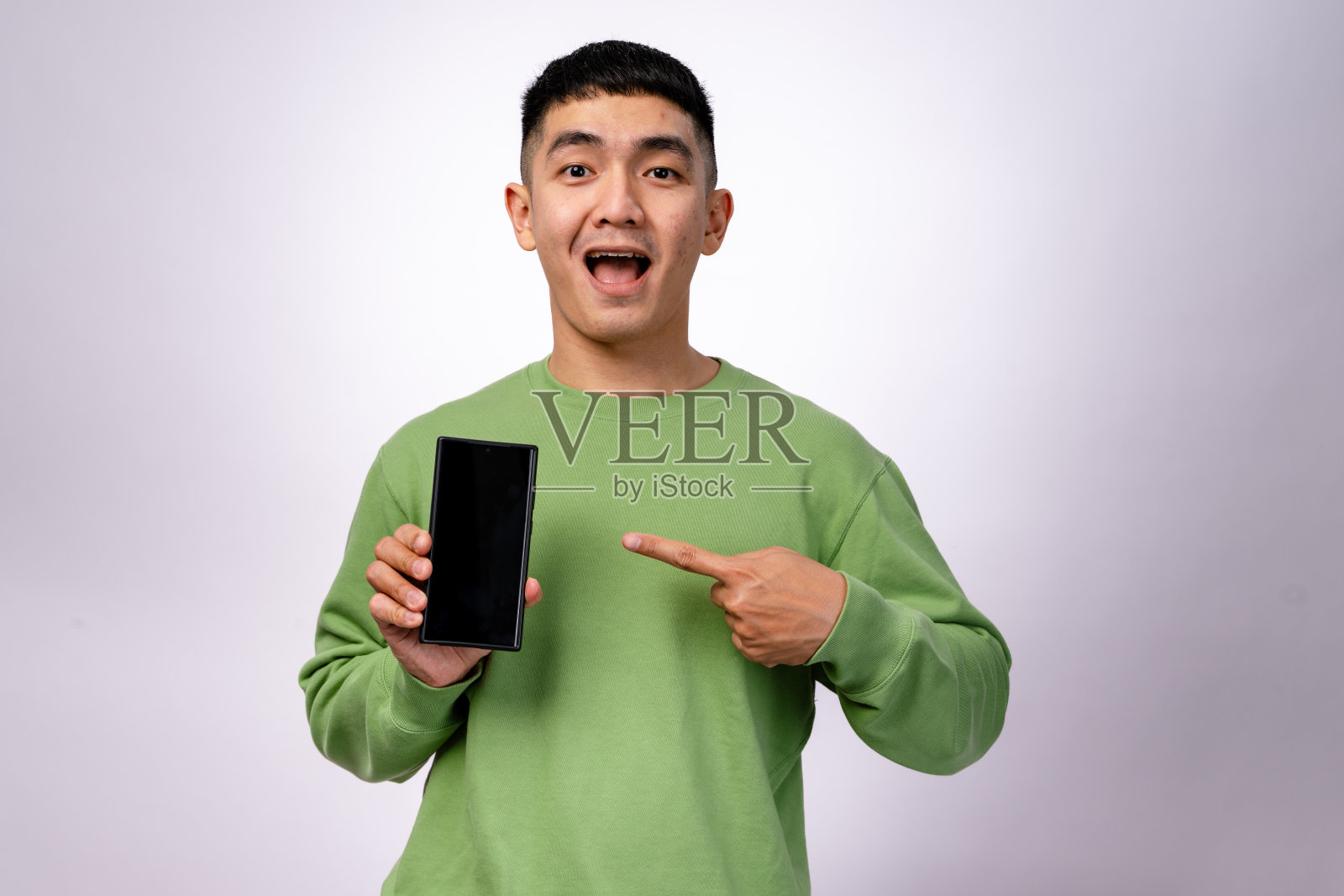 一位穿着绿色卫衣的亚洲男子微笑着,指着一部空白屏幕的智能手机。他用一只手握着手机,兴奋地朝它做手势。背景是白色的。照片摄影图片