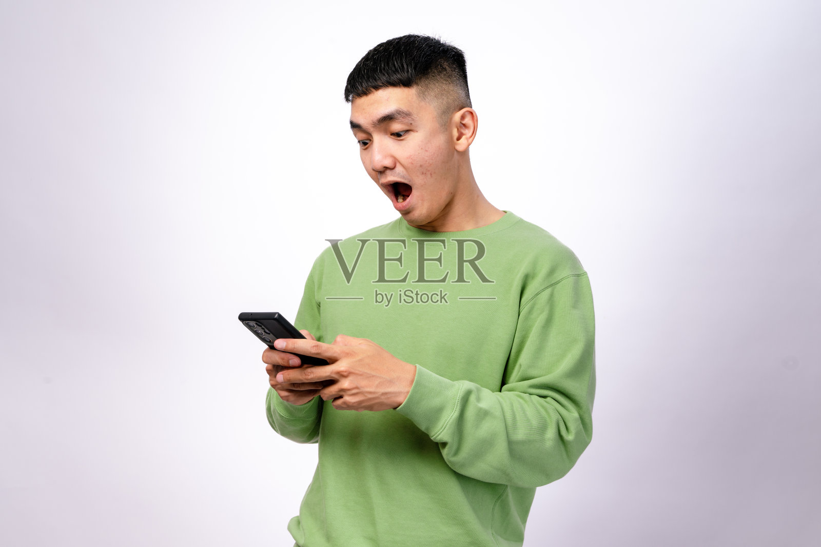 一位穿着绿色卫衣的亚洲男子专注于他的智能手机,双手握住手机,目光注视着屏幕。他的表情中性。背景是白色的。照片摄影图片