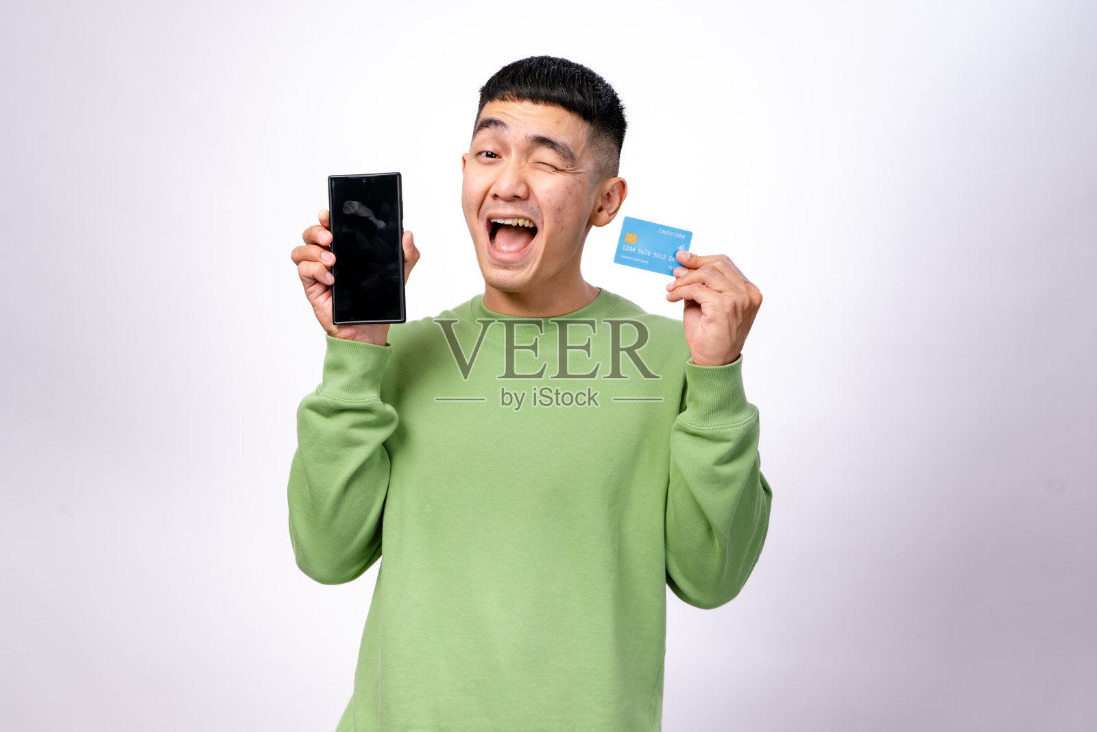 一位兴奋的亚洲男子穿着绿色卫衣,一只手拿着智能手机,另一只手拿着蓝色信用卡,朝着镜头展示。他看起来很开心。背景是白色的。照片摄影图片